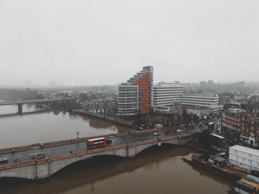 Aerial view of a cityscape showing the Thames River with two bridges crossing over it, including a road with moving vehicles such as buses and cars. On the near side of the river, there is a modern mu