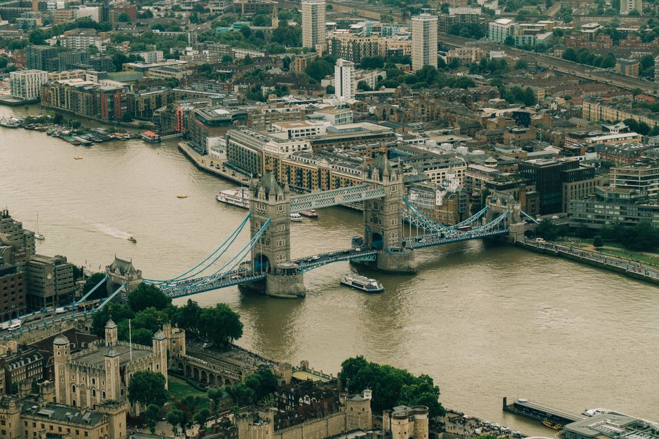 Aerial view of a cityscape showing the Thames River with two bridges crossing over it, including a road with moving vehicles such as buses and cars. On the near side of the river, there is a modern mu